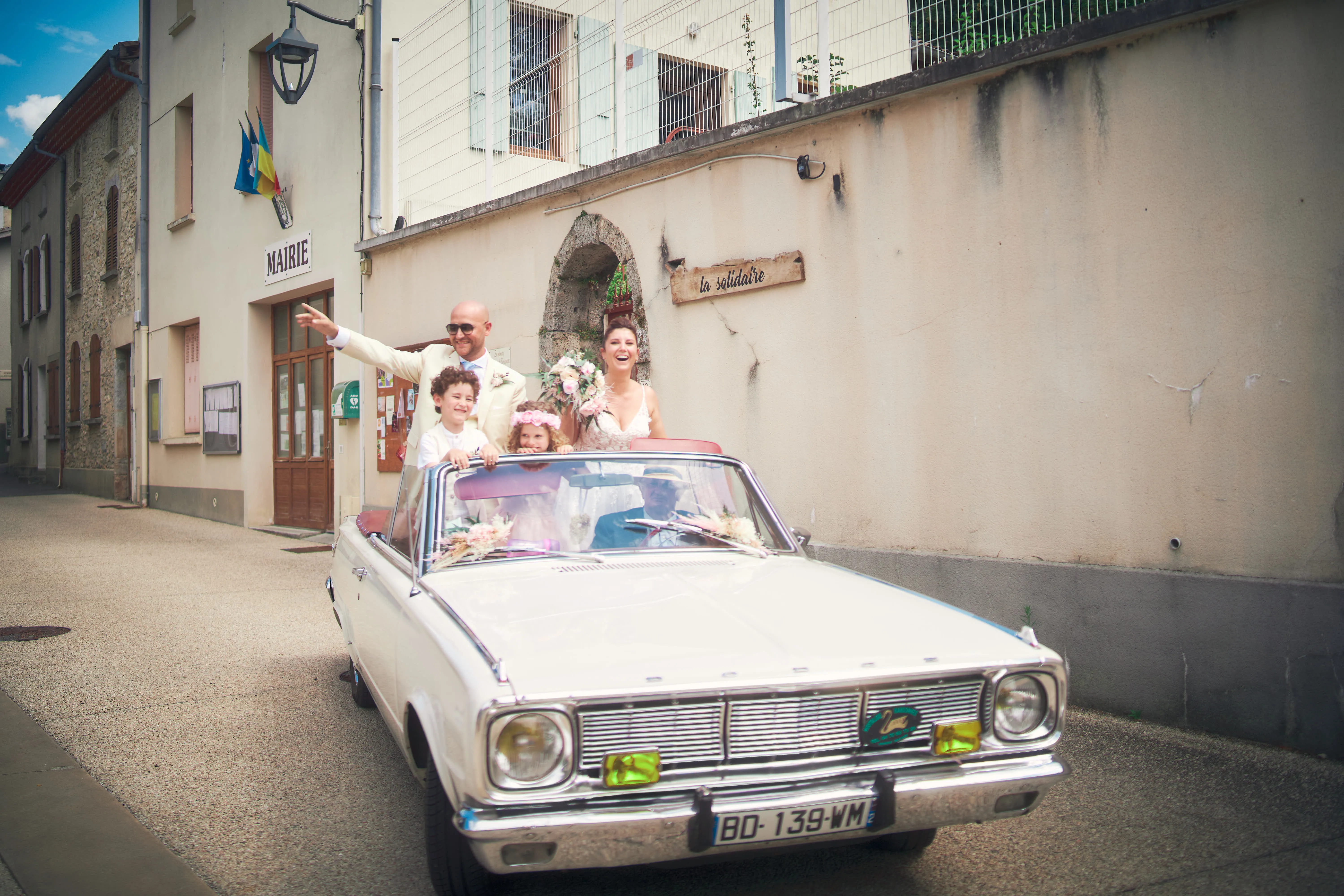 Mariés saluant depuis une voiture ancienne dans une rue de village.