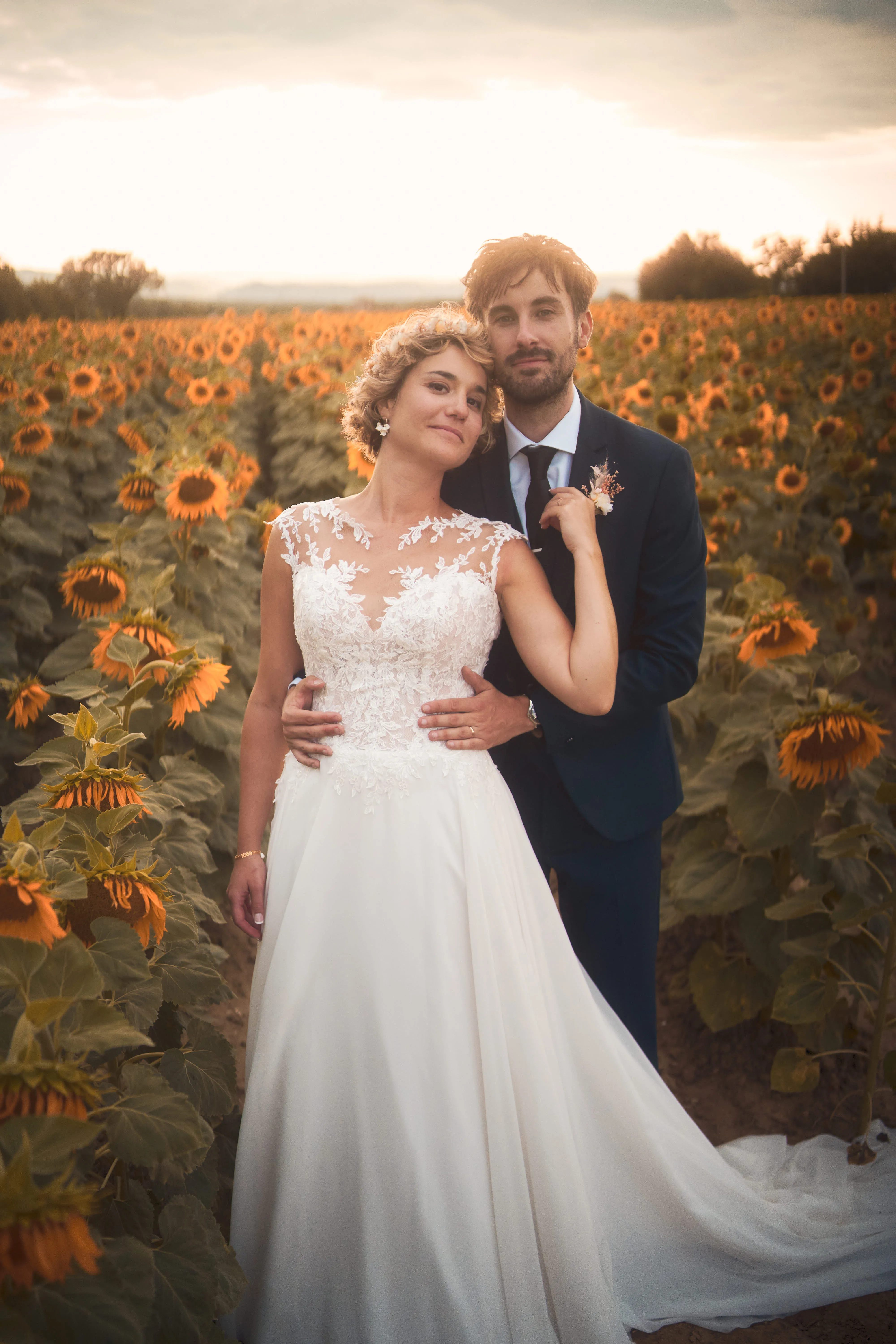 Mariés enlacés devant un champ de tournesols au coucher du soleil.